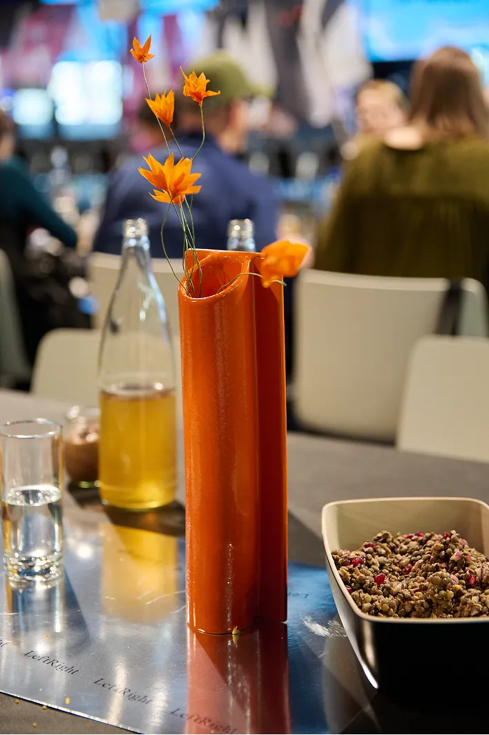 Vase orange aux fleurs éclatantes sur une surface réfléchissante, à côté d'un verre de liquide jaune et d'un bol de graines. Arrière-plan flou de personnes assises.