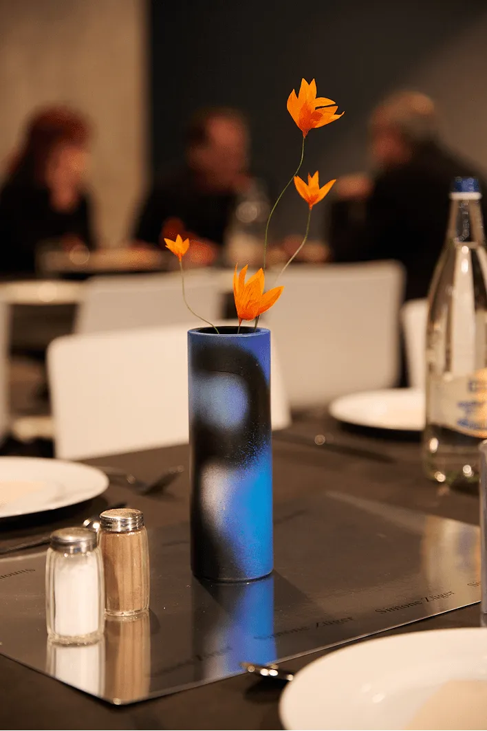 Un vase bleu dégradé, rempli de fleurs orange, trône sur une table de restaurant, à côté des salières et poivrières. En arrière-plan, des clients aux silhouettes floues conversent, créant une ambiance chaleureuse et intime.
