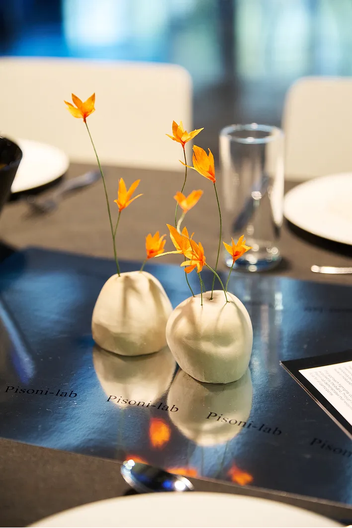 Deux petits vases blancs remplis de fleurs orange ornent une table aux reflets chatoyants, entourée de couverts. L'atmosphère est à la fois élégante et paisible.